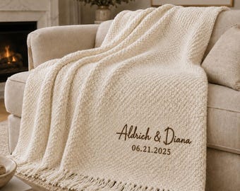 May include: A cream-colored knitted throw blanket with fringe trim, featuring brown embroidered text that reads Aldrich & Diana and the date 06.21.2025. The blanket has a textured, woven pattern and is draped over a light-colored upholstered sofa.