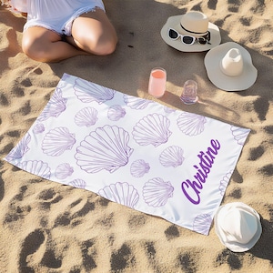 May include: White beach towel with a purple seashell pattern and the name "Christine" in purple script. The towel is rectangular and lies on a sandy surface, with beach hats and drinks nearby. A beach accessory.