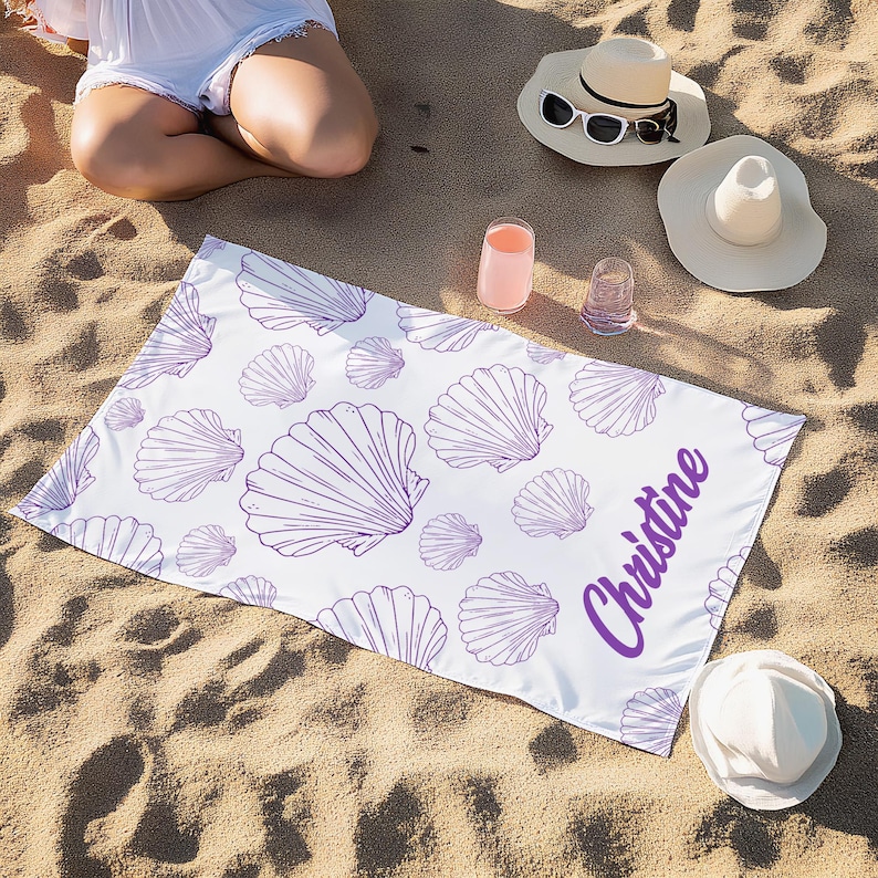May include: White beach towel with a purple seashell pattern and the name "Christine" in purple script. The towel is rectangular and lies on a sandy surface, with beach hats and drinks nearby. A beach accessory.