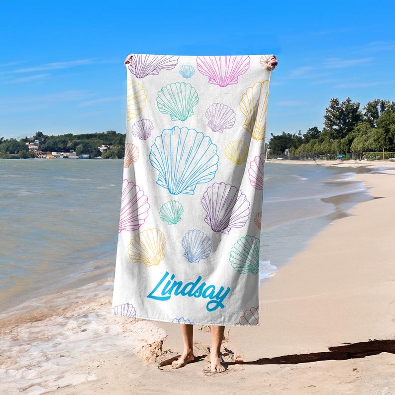 May include: White beach towel with a pattern of colorful seashells in shades of blue, pink, yellow, and green. The word "Lindsay" is written in a teal script font across the bottom of the towel. The towel is held up on a beach.