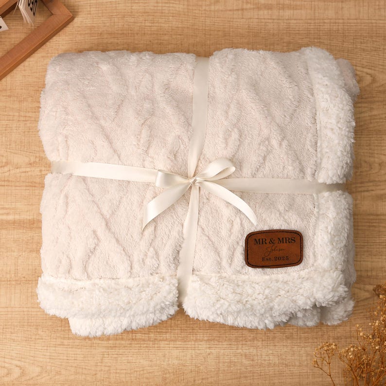 May include: Cream-colored, plush blanket with a cable knit design, wrapped with a white satin ribbon tied in a bow. A brown leather patch with "MR & MRS" and "Est. 2025" is attached. The blanket has a soft, fluffy texture.