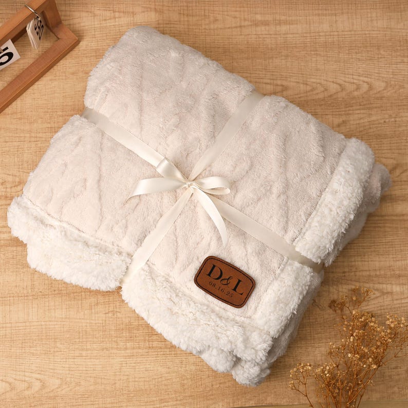 May include: A cream-colored, plush blanket with a cable knit design, tied with a white ribbon. A brown leather patch with the initials "D&L" and the date "08.16.25" is sewn on the blanket. The blanket is folded and ready for gifting.