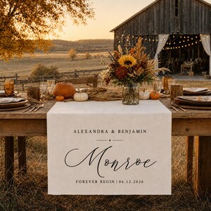 May include: A white fabric table runner featuring the printed text ALEXANDRA & BENJAMIN, the surname Monroe in a large script font, and the date 06.12.2026. The runner is draped over a wooden table set with pumpkins and a floral centerpiece.