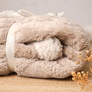 May include: A rolled-up, plush blanket in shades of beige and cream, tied with a cream-colored ribbon. The blanket has a soft, textured appearance, suggesting a cozy and comfortable feel. The overall aesthetic is neutral and inviting.