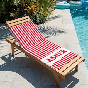 May include: Wooden outdoor chaise lounge with a red and white striped towel. The towel has the name "ASHER" printed in red block letters. The lounge is positioned next to a swimming pool.