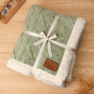 May include: A folded sage green blanket with a cable knit design, trimmed with white sherpa fleece. The blanket is tied with a white ribbon and bow, and features a brown leather tag that reads "The Bowman Family."