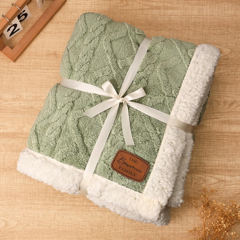 May include: A folded sage green blanket with a cable knit design, trimmed with white sherpa fleece. The blanket is tied with a white ribbon and bow, and features a brown leather tag that reads "The Bowman Family."