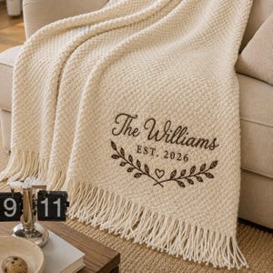 Può includere: Una coperta testurizzata color crema con bordi sfrangiati, caratterizzata da un ricamo marrone scuro che recita The Williams EST. 2026 sopra un motivo decorativo a ramo e cuore. Il tessuto presenta un motivo a maglia in rilievo su tutta la superficie.