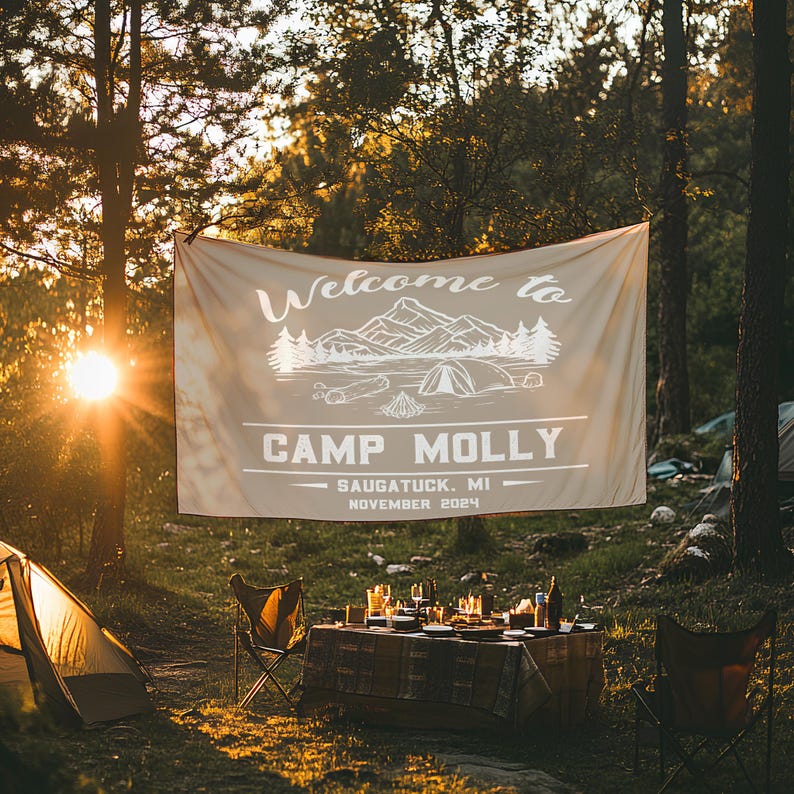 May include: A beige canvas banner with white text and graphics reads "Welcome to Camp Molly - Saugatuck, MI - November 2024". The banner is hanging between two trees in a forest setting. A table with chairs and dishes is set up in front of the banner.