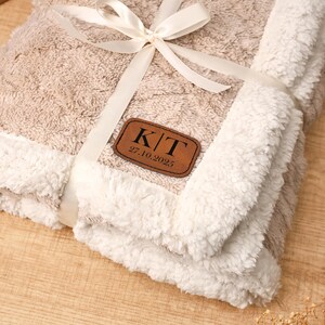 May include: A folded beige and white blanket tied with a cream ribbon. The blanket has a textured pattern and a white, fluffy border. A brown leather patch with the initials "K/T" and the date "27.10.2025" is attached to the blanket.