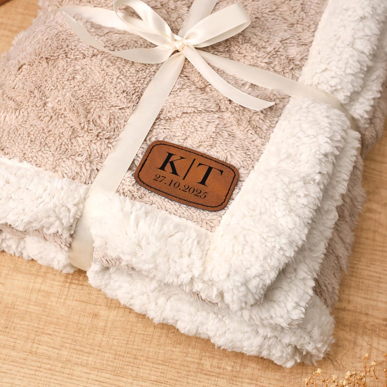 May include: A folded beige and white blanket tied with a cream ribbon. The blanket has a textured pattern and a white, fluffy border. A brown leather patch with the initials "K/T" and the date "27.10.2025" is attached to the blanket.