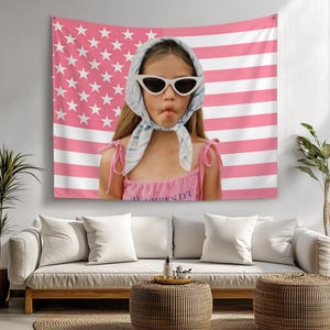 Puede incluir: Un tapiz con la bandera estadounidense rosa y blanca cuelga detrás de una persona con gafas de sol y un pañuelo en la cabeza. La persona lleva una camiseta rosa con el texto "MELAIRES D'U". Un sofá blanco y una mesa tejida están en primer plano.