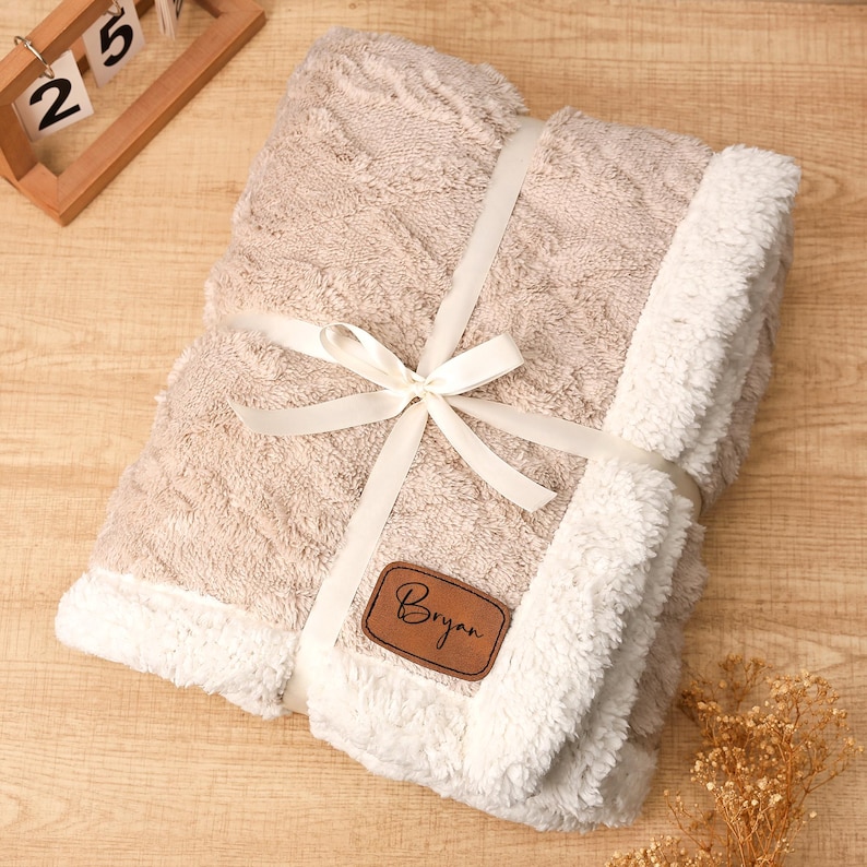 May include: A beige blanket with a diamond pattern and white sherpa trim, tied with a white ribbon. A brown leather patch with the name "Bryan" is attached. The blanket is folded and ready to be gifted.