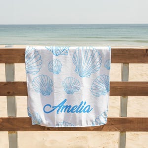May include: White beach towel with a blue seashell pattern and the name "Amelia" in blue script. The towel is hanging on a wooden fence with a beach and ocean in the background.