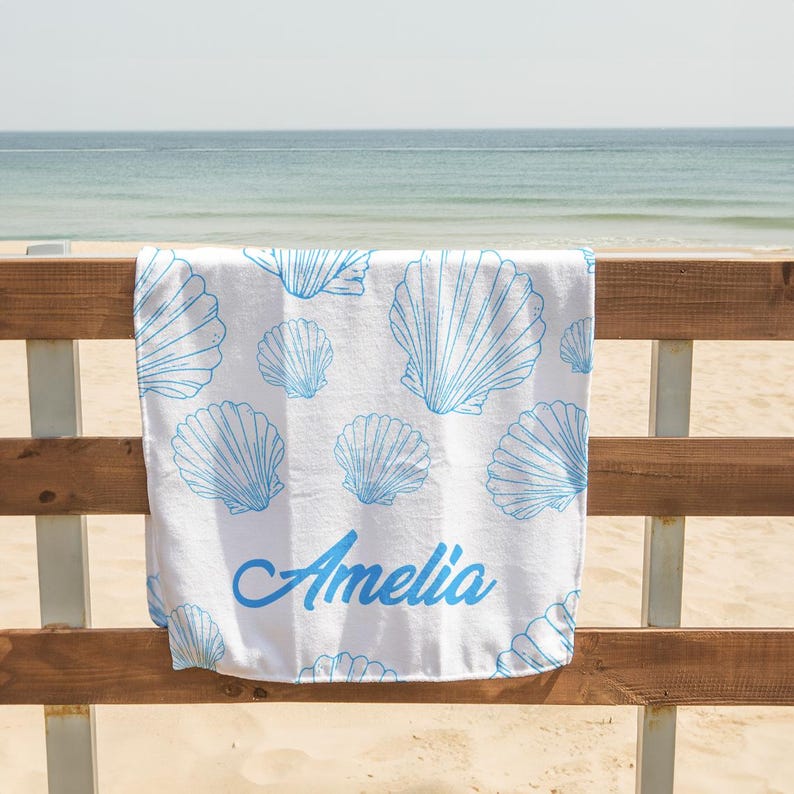 May include: White beach towel with a blue seashell pattern and the name "Amelia" in blue script. The towel is hanging on a wooden fence with a beach and ocean in the background.