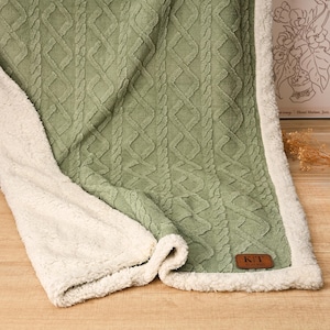 May include: Sage green and cream-colored blanket with a cable knit design. The blanket has a soft, plush cream-colored sherpa lining. A brown leather tag with the initials "K/T" is sewn on the blanket.