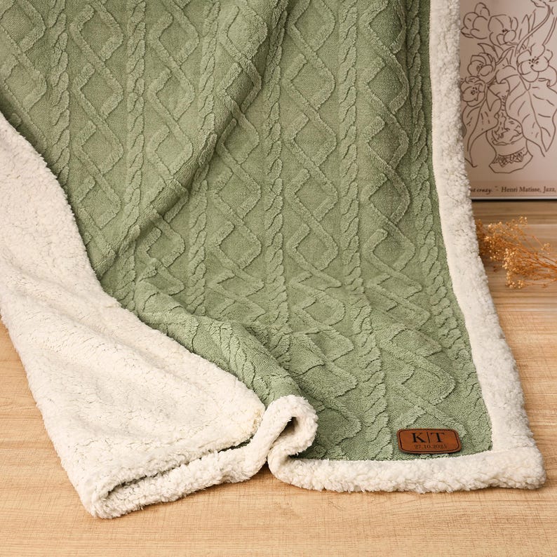 May include: Sage green and cream-colored blanket with a cable knit design. The blanket has a soft, plush cream-colored sherpa lining. A brown leather tag with the initials "K/T" is sewn on the blanket.