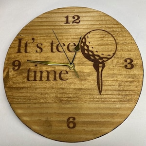 Puede incluir: Un reloj de madera con un diseño de bola de golf. La esfera del reloj tiene los números 12, 3, 6 y 9. Las manecillas del reloj son doradas. La esfera del reloj también tiene el texto "It's tee time".