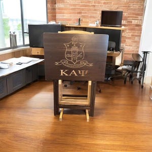 May include: A dark brown wooden folding table with a light brown wooden frame. The table top has a laser engraved design of a crest with the letters "ΚΑΨ" below it.