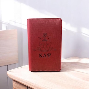 May include: A red leather wallet with a black Greek fraternity crest and the letters "ΚΑΨ" engraved on the front.