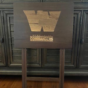 Puede incluir: Una bandeja de TV de madera marrón oscuro con el logotipo de los Washington Commanders. La bandeja tiene un logotipo "W" de vetas de madera más claras y el nombre del equipo en un recuadro rectangular. La bandeja está plegada.