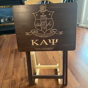 May include: A dark brown wooden serving tray with a gold engraved fraternity crest and the letters "ΚΑΨ" and the text "LIFE MEMBER". The tray is on a wooden folding stand.