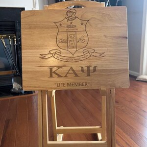 May include: A wooden serving tray with a folding stand. The tray features an engraved crest with the letters "ΚΑΨ" and the text "Life Member".