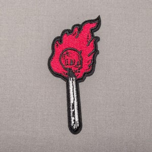 May include: An embroidered patch featuring a lit matchstick with a bright red flame. The flame contains a subtle skull design in the center. The matchstick has a white shaft with black shading and a thick black border outlining the entire shape.