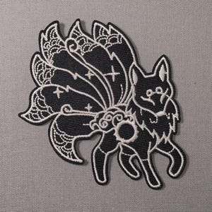 May include: A black and white embroidered patch featuring a stylized nine-tailed fox. The design includes intricate white line work detailing the multiple tails with decorative patterns, small star accents, and a circular motif on the fox's body.