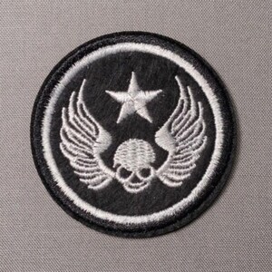 May include: A circular embroidered patch featuring a black background with a white border. The design includes a central white skull with detailed wings extending outward on both sides and a single five-pointed white star positioned above.