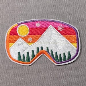 May include: An oval-shaped embroidered patch featuring a mountain landscape with white snow-capped peaks, a yellow sun, and green pine trees against a sunset sky of pink, orange, and yellow stripes, accented with small white embroidered snowflakes.