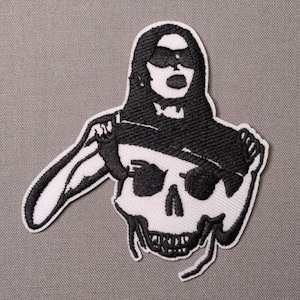 May include: An embroidered patch featuring a black and white design of a figure with long hair wearing a mask over their eyes, positioned above a skull. The figure holds a whip-like cord that extends to the side of the skull graphic.