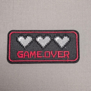 May include: A rectangular embroidered patch with a black background and a red border. The design features three pixelated light gray hearts arranged horizontally above the red pixelated text that reads GAME OVER in all capital letters.