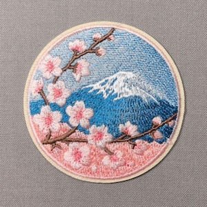 May include: A circular embroidered patch featuring a snow-capped mountain peak in deep blue against a lighter blue sky. Brown branches with light pink cherry blossoms extend across the foreground, framing the mountain scene with a cream border.