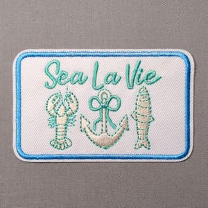 May include: A rectangular embroidered patch with a white background and a blue border. It features the teal text Sea La Vie above a centered anchor, a lobster on the left, and a fish on the right, all stitched in teal and light tan thread.