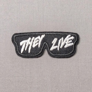 May include: A black embroidered patch shaped like a pair of sunglasses with the white text THEY LIVE stitched across the lenses. The patch features a dark gray border and is set against a light gray fabric background with a visible woven texture.