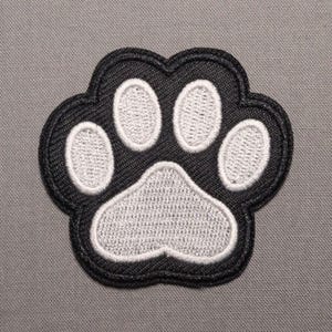 May include: A black and white embroidered patch in the shape of a paw print. The design features a large central pad and four smaller toe pads, all outlined in black thread against a textured fabric background with a stitched border.