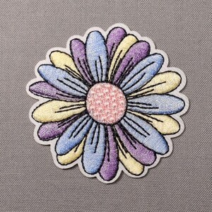 May include: A circular embroidered patch featuring a flower design with a textured pink center. The petals are arranged in alternating colors of light blue, pale yellow, and purple, each outlined with dark thread on a white fabric border.