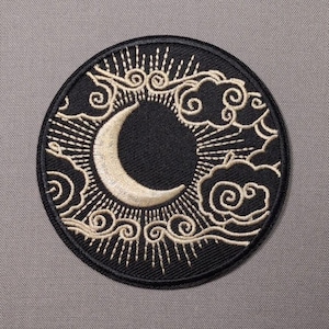 May include: A circular embroidered patch featuring a black background with a central crescent moon in cream thread. The design includes radiating sunburst lines and stylized swirling clouds in cream, all enclosed by a black stitched border.
