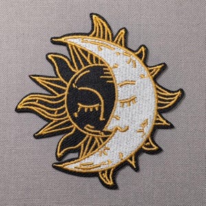 May include: An embroidered patch featuring a sun and moon design. The sun is black with gold rays, while the crescent moon is white with gold accents. Both celestial bodies have closed eyes and eyelashes stitched in gold thread on a textured fabric.