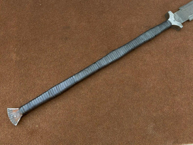 May include: A long, black-wrapped sword with a silver blade and a triangular handle. The sword is lying on a brown surface.