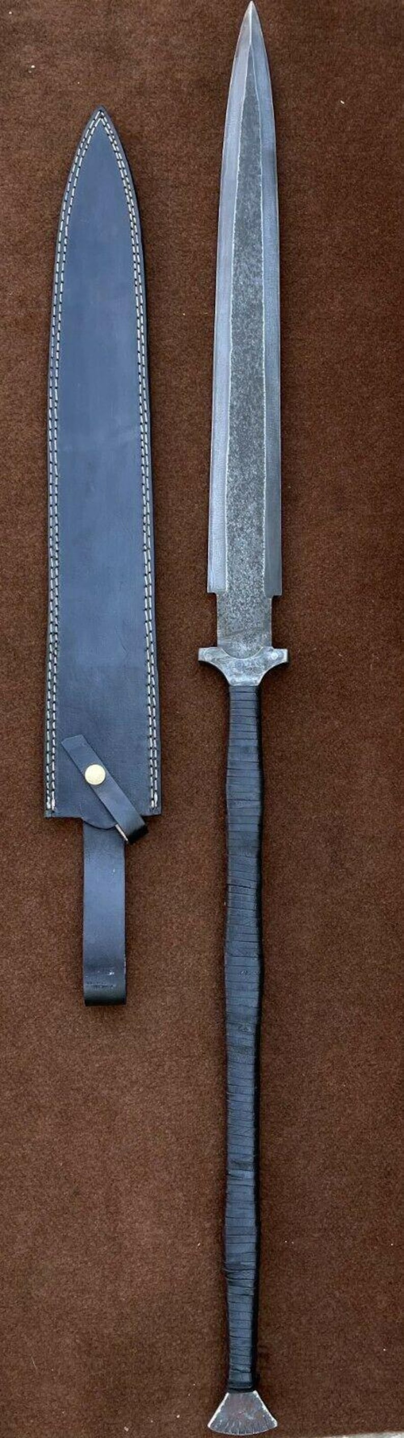 May include: A long, silver-colored sword with a black leather handle and a matching black leather sheath. The sword is on a brown background.