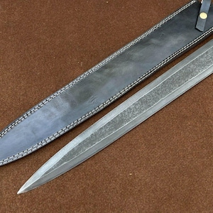 May include: A long, silver-colored knife with a black leather sheath. The knife has a textured blade and a simple handle.