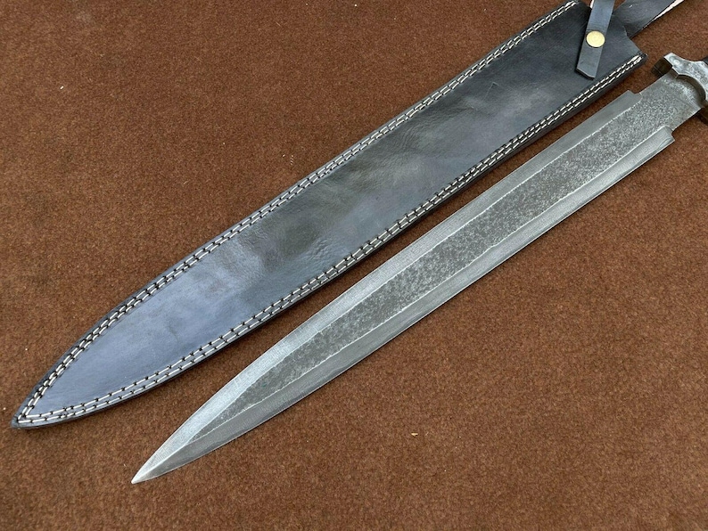 May include: A long, silver-colored knife with a black leather sheath. The knife has a textured blade and a simple handle.