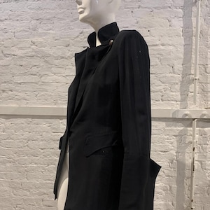 Peut inclure: Un blazer noir avec un col montant et une fermeture à bouton unique. Le blazer a des manches longues et une coupe ajustée, avec une poche sur le devant. Le vêtement est présenté sur un mannequin devant un mur de briques blanches.