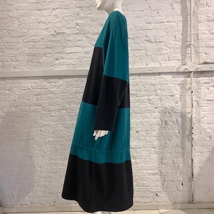 May include: A long-sleeved midi dress featuring a color-blocked pattern of horizontal teal and black stripes. The garment has a round neckline and a gathered waistline detail, creating a relaxed silhouette that falls below the knee in length.