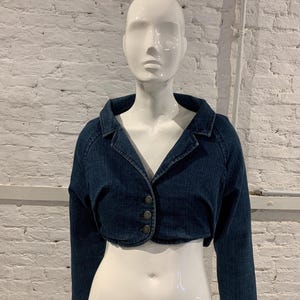 Pode incluir: Casaco curto de ganga azul escuro. Apresenta uma gola clássica, mangas compridas e três botões escuros. O casaco é exibido num manequim branco contra uma parede de tijolos brancos. O casaco tem cerca de 30 cm de comprimento.