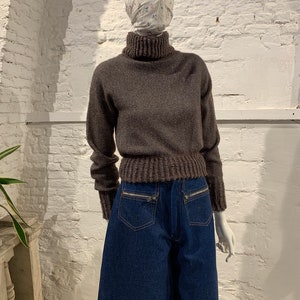 Op de afbeelding: Een bruine trui met een hoge hals en een geribbelde textuur en een donkerblauwe spijkerbroek met ritszakken.