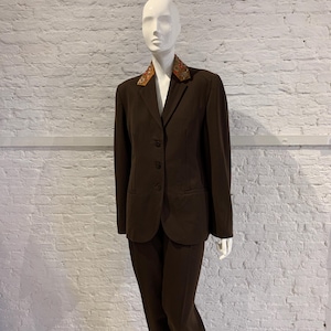 Può includere: Un completo marrone su un manichino. La giacca ha un colletto fantasia e tre bottoni, abbinata a pantaloni coordinati. L'abito è su misura. Lo sfondo è un muro di mattoni bianchi. L'abito è uno stile classico.