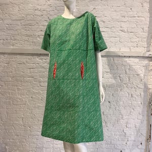 Può includere: Un abito verde al ginocchio con un motivo a linee ondulate bianche, maniche corte e scollo rotondo. Il capo presenta due tasche verticali a fessura sul davanti, ciascuna accentuata da un piccolo inserto in tessuto a fiori rosso.